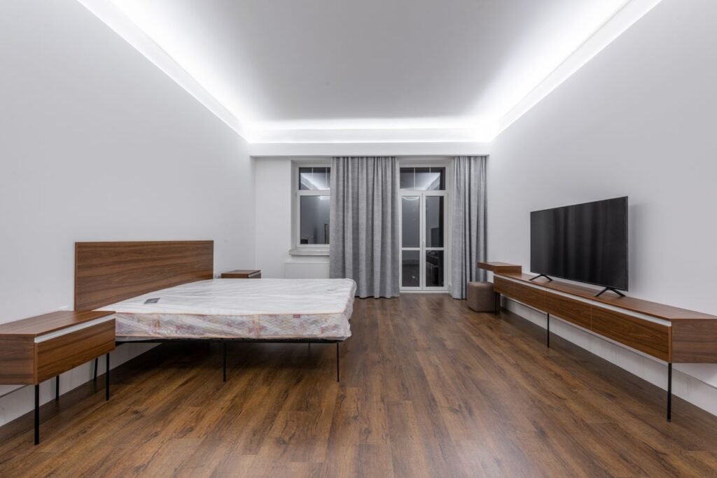 Bed with mattress placed against wall with TV on wooden shelf in spacious bedroom with window and door at home