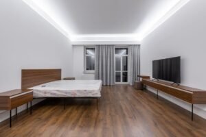 Bed with mattress placed against wall with TV on wooden shelf in spacious bedroom with window and door at home
